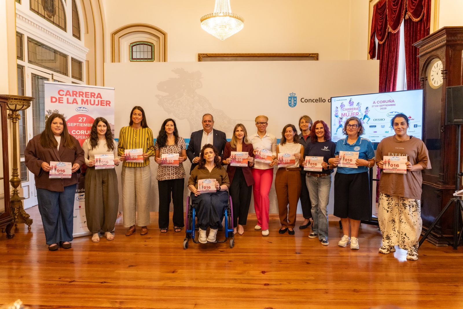 Grupo de personas con dorsales de la Carrera de la Mujer, una de ellas en silla de ruedas, posan en un salón con suelo de madera junto a un cartel de la carrera