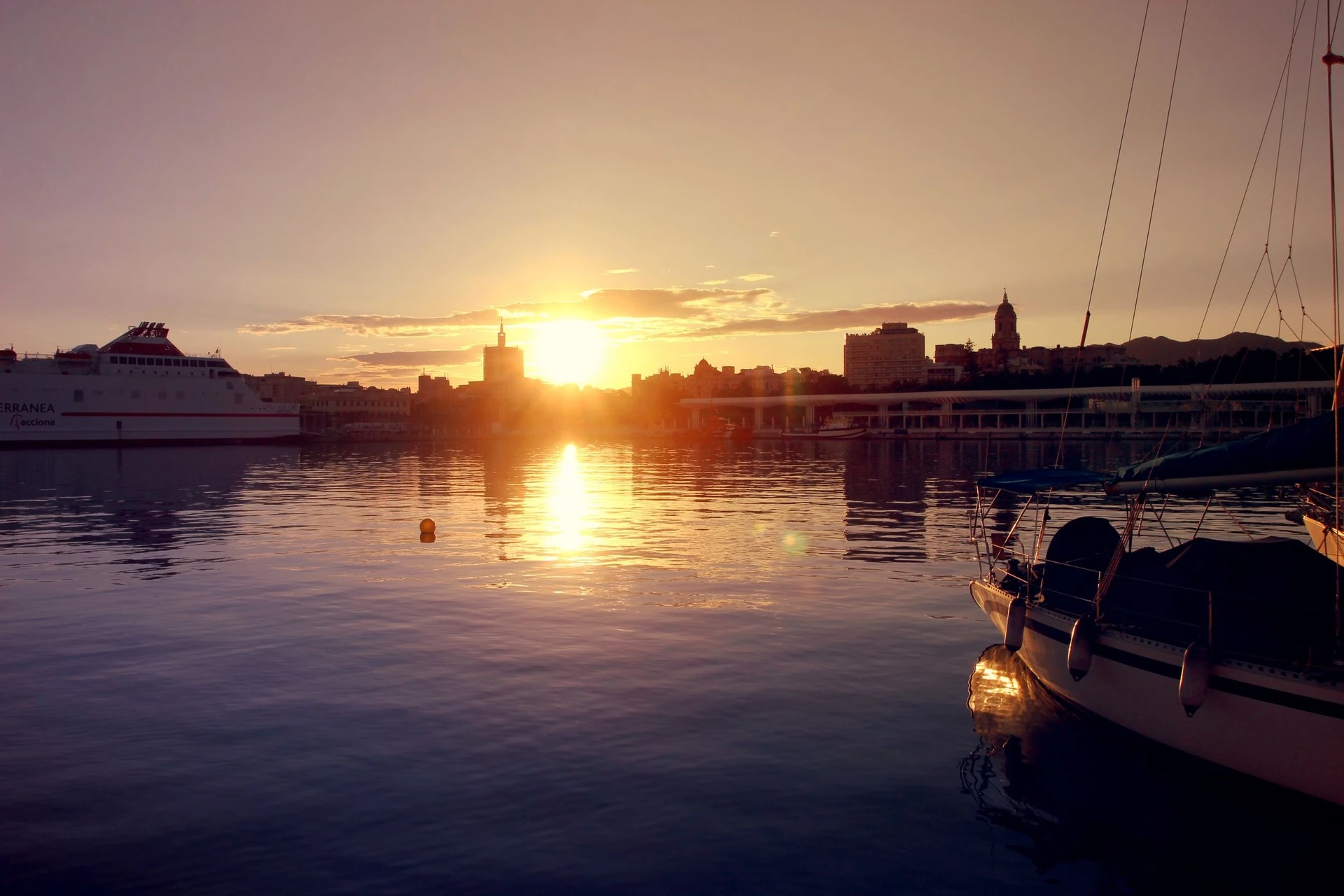 Puerto de Málaga al atardecer, con el sol sobre el horizonte y veleros en primer plano