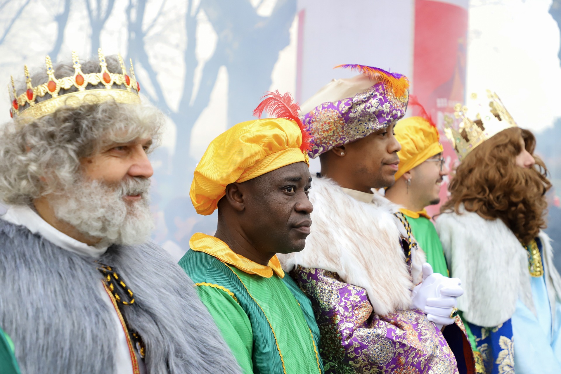 Los Reyes Magos y sus pajes, de perfil, con coronas, turbantes de plumas y capas de piel, al aire libre entre árboles.