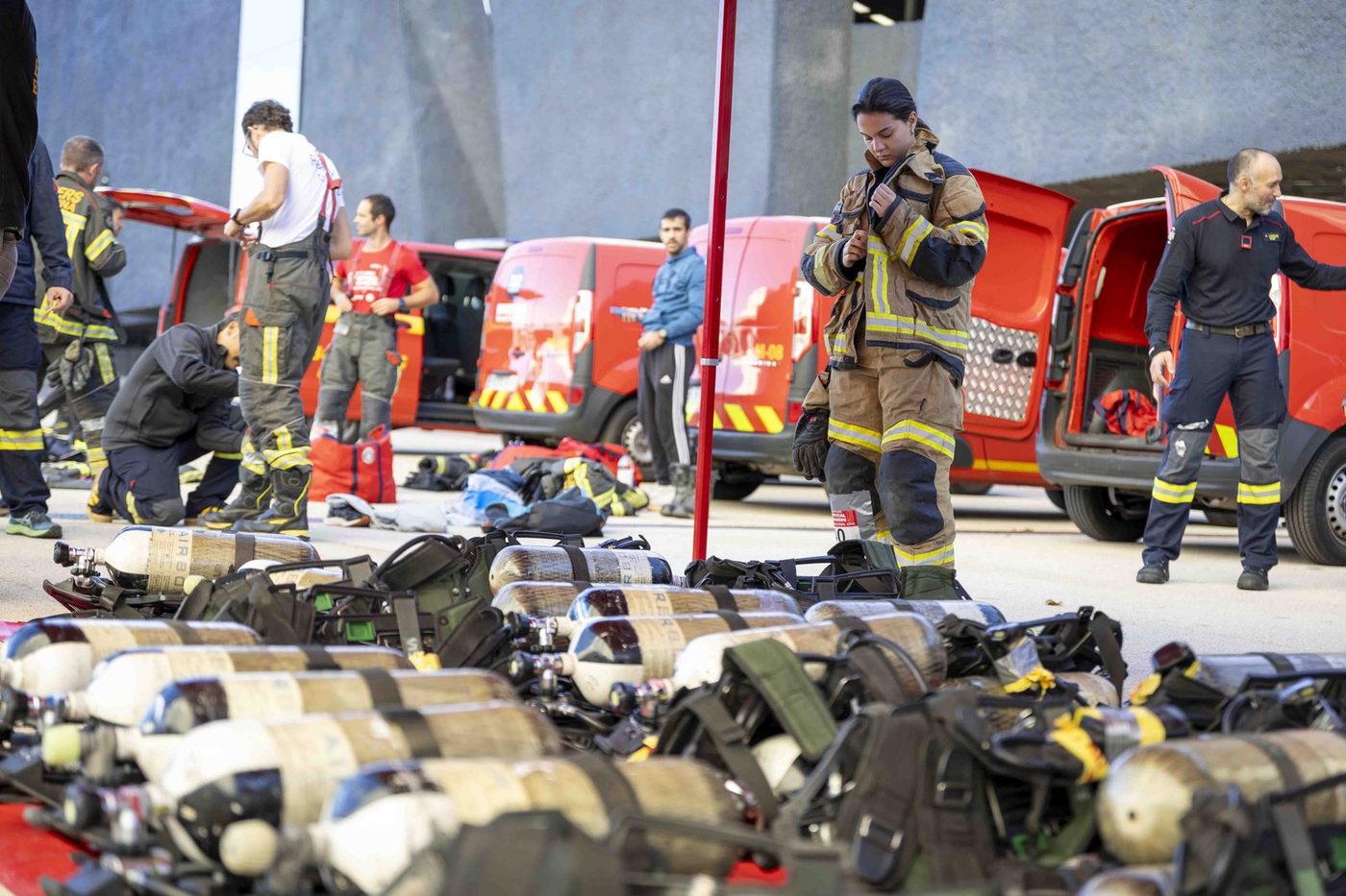Bomberas y bomberos preparan su equipo antes de la salida, con las botellas de aire en primer termino y los vehiculos de Bombers de Barcelona al fondo.
