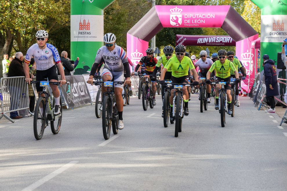 Ciclistas de montaña saliendo por el asfalto tras pasar bajo el arco hinchable rosa de la Diputación de León, con vallas y público a los lados