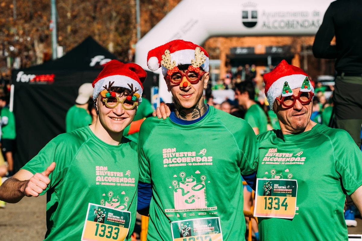 Tres corredores con la camiseta verde de la San Silvestre de Alcobendas, gorros de Papá Noel y gafas de fiesta, sonríen junto al arco del Ayuntamiento de Alcobendas